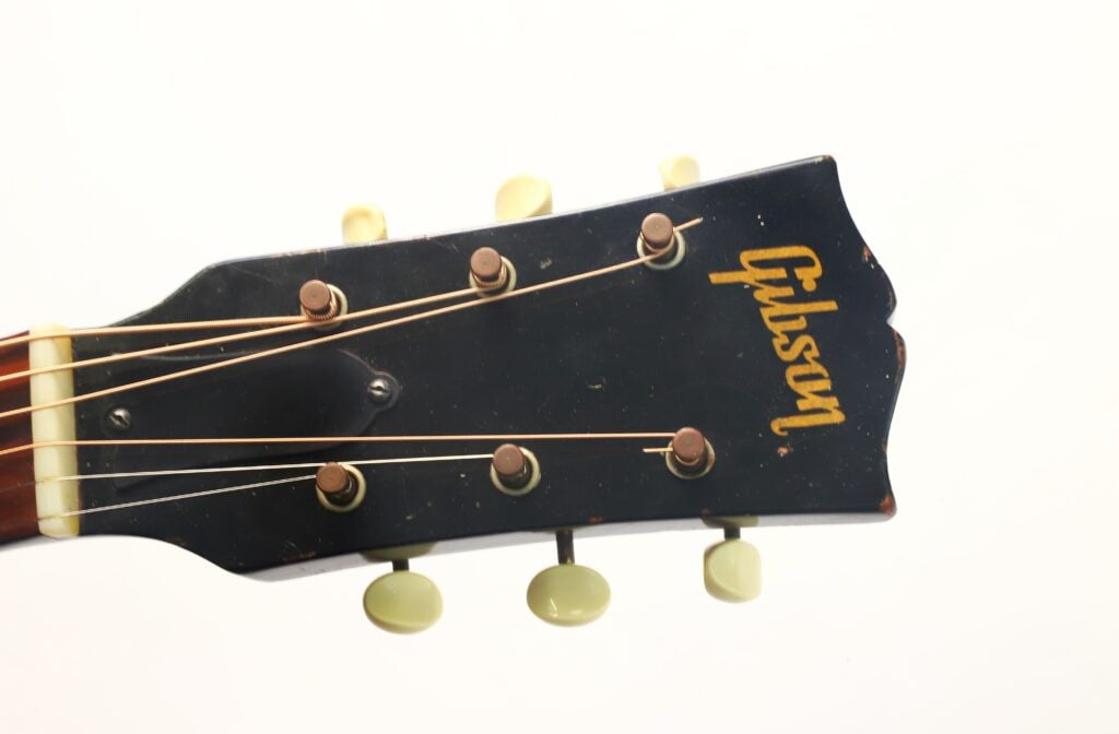 Nahaufnahme des Kopfes einer schwarzen Gibson L-48 Akustikgitarre aus dem Jahr 1948, auf der die Stimmwirbel und Saiten zu sehen sind, fotografiert vor einem schlichten weißen Hintergrund. Nahaufnahme des Kopfes einer schwarzen Gibson L-48 Akustikgitarre aus dem Jahr 1948, auf der die Stimmwirbel und Saiten zu sehen sind, fotografiert vor einem schlichten weißen Hintergrund.