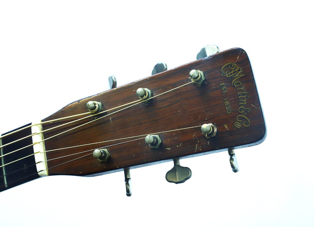 Nahaufnahme des Kopfes einer Martin 000-18 Akustikgitarre aus dem Jahr 1960, mit Stimmwirbeln und einem goldenen Logo auf Holz vor einem weißen Hintergrund. Nahaufnahme des Kopfes einer Martin 000-18 Akustikgitarre aus dem Jahr 1960, mit Stimmwirbeln und einem goldenen Logo auf Holz vor einem weißen Hintergrund.
