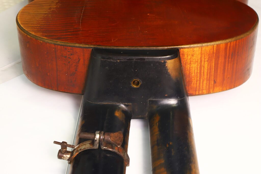 Eine Nahaufnahme der Rückseite einer Kontragitarre vom Typ „Stil 1900“ von Gustav Keller Stauffer, auf deren Hals auf einem weißen Untergrund eine schwarze Metallvorrichtung befestigt ist. Eine Nahaufnahme der Rückseite einer Kontragitarre vom Typ „Stil 1900“ von Gustav Keller Stauffer, auf deren Hals auf einem weißen Untergrund eine schwarze Metallvorrichtung befestigt ist.