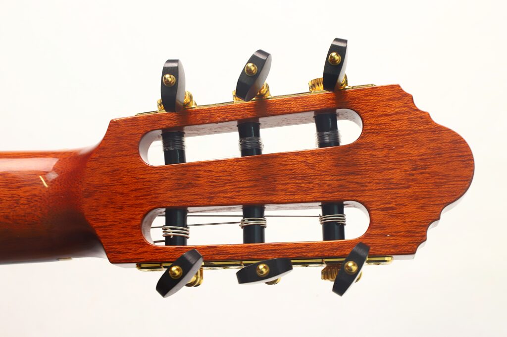 Nahaufnahme des Kopfstücks einer „Antonius Müller 2012 Doubletop Klassische Gitarre“ mit sechs Stimmwirbeln aus schwarz-goldenem Metall und Nylonsaiten vor einem schlichten weißen Hintergrund. Nahaufnahme des Kopfstücks einer „Antonius Müller 2012 Doubletop Klassische Gitarre“ mit sechs Stimmwirbeln aus schwarz-goldenem Metall und Nylonsaiten vor einem schlichten weißen Hintergrund.