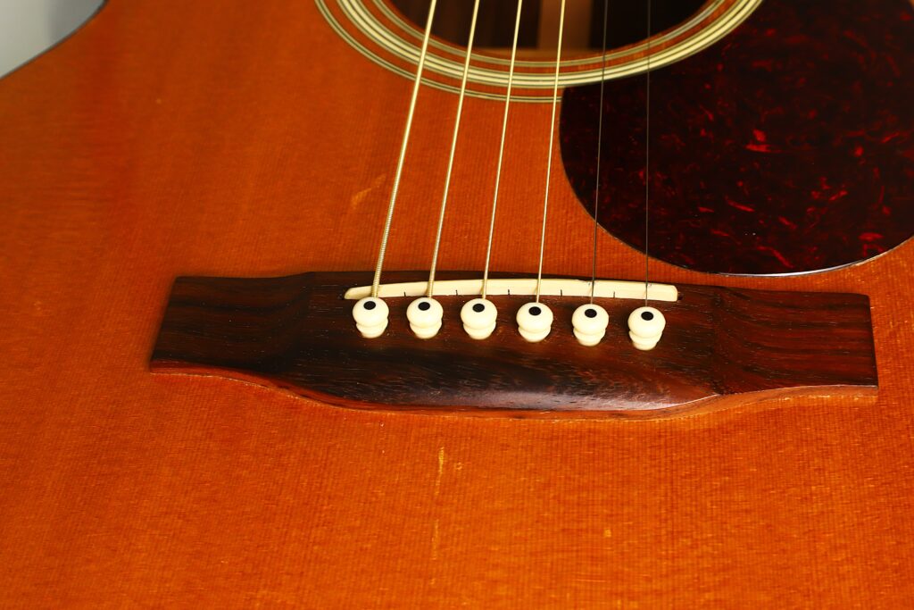 Nahaufnahme des Stegs und der Saiten einer MARTIN OM-21 Akustikgitarre aus dem Jahr 1995, auf der sechs Stegstifte, der untere Teil des Schalllochs und ein braunes Schlagbrett auf dem Holzkorpus zu sehen sind. Nahaufnahme des Stegs und der Saiten einer MARTIN OM-21 Akustikgitarre aus dem Jahr 1995, auf der sechs Stegstifte, der untere Teil des Schalllochs und ein braunes Schlagbrett auf dem Holzkorpus zu sehen sind.