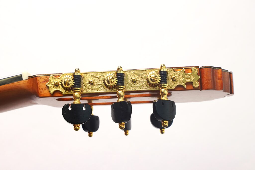Nahaufnahme der Stimmwirbel und der kunstvollen Vergoldung am Kopf einer Gerhard Arns 2026 Smallman Klassische Gitarre, horizontal vor einem schlichten weißen Hintergrund abgebildet. Nahaufnahme der Stimmwirbel und der kunstvollen Vergoldung am Kopf einer Gerhard Arns 2026 Smallman Klassische Gitarre, horizontal vor einem schlichten weißen Hintergrund abgebildet.