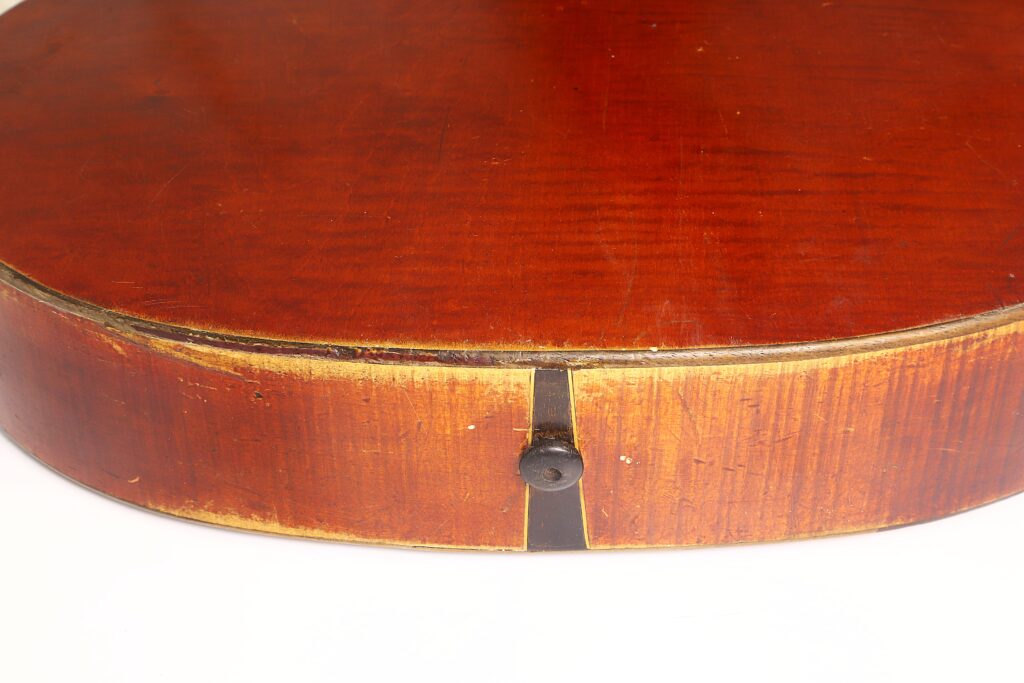 Nahaufnahme der Unterkante einer Kontragitarre vom Typ „Stil 1900“ von Gustav Keller Stauffer, die einen dunklen Knopf sowie abgenutztes, strukturiertes Holz mit glänzender Oberfläche zeigt. Nahaufnahme der Unterkante einer Kontragitarre vom Typ „Stil 1900“ von Gustav Keller Stauffer, die einen dunklen Knopf sowie abgenutztes, strukturiertes Holz mit glänzender Oberfläche zeigt.