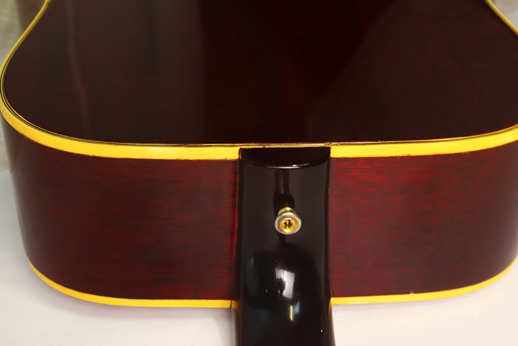 Nahaufnahme der Rückseite der Gibson SJN 1968 Akustikgitarre, die die glänzende, dunkle Holzoberfläche, die gelbe Einfassung und den Halsansatz mit einem Gurtknopf hervorhebt. Nahaufnahme der Rückseite der Gibson SJN 1968 Akustikgitarre, die die glänzende, dunkle Holzoberfläche, die gelbe Einfassung und den Halsansatz mit einem Gurtknopf hervorhebt.