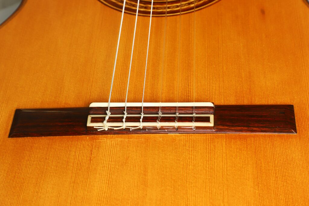 Nahaufnahme des Holzstegs und der Nylonsaiten der „Friederich Klassische Gitarre“ von Stefan Nitschke aus dem Jahr 2015 mit hellbraunem Korpus. Der rechteckige Steg hält die Saiten fest, die an der Unterseite verknotet sind. Nahaufnahme des Holzstegs und der Nylonsaiten der „Friederich Klassische Gitarre“ von Stefan Nitschke aus dem Jahr 2015 mit hellbraunem Korpus. Der rechteckige Steg hält die Saiten fest, die an der Unterseite verknotet sind.