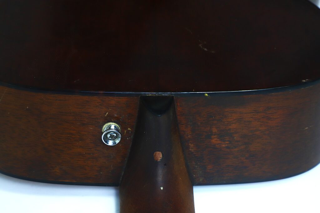 Nahaufnahme der Rückseite im Übergang vom Hals zum Korpus einer Martin 000-18 Akustikgitarre aus dem Jahr 1960, wobei die braune Holzoberfläche und ein Metall-Gurtknopf an der Seite besonders hervorgehoben werden. Nahaufnahme der Rückseite im Übergang vom Hals zum Korpus einer Martin 000-18 Akustikgitarre aus dem Jahr 1960, wobei die braune Holzoberfläche und ein Metall-Gurtknopf an der Seite besonders hervorgehoben werden.