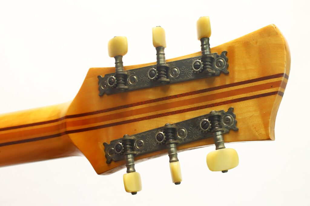 Nahaufnahme der Rückseite des hölzernen Kopfes einer Artur-Lang-Jazzgitarre aus den 1950er Jahren, die sechs Stimmwirbel mit cremefarbenen Knöpfen und dekorativen Metallplättchen vor einem schlichten weißen Hintergrund zeigt. Nahaufnahme der Rückseite des hölzernen Kopfes einer Artur-Lang-Jazzgitarre aus den 1950er Jahren, die sechs Stimmwirbel mit cremefarbenen Knöpfen und dekorativen Metallplättchen vor einem schlichten weißen Hintergrund zeigt.