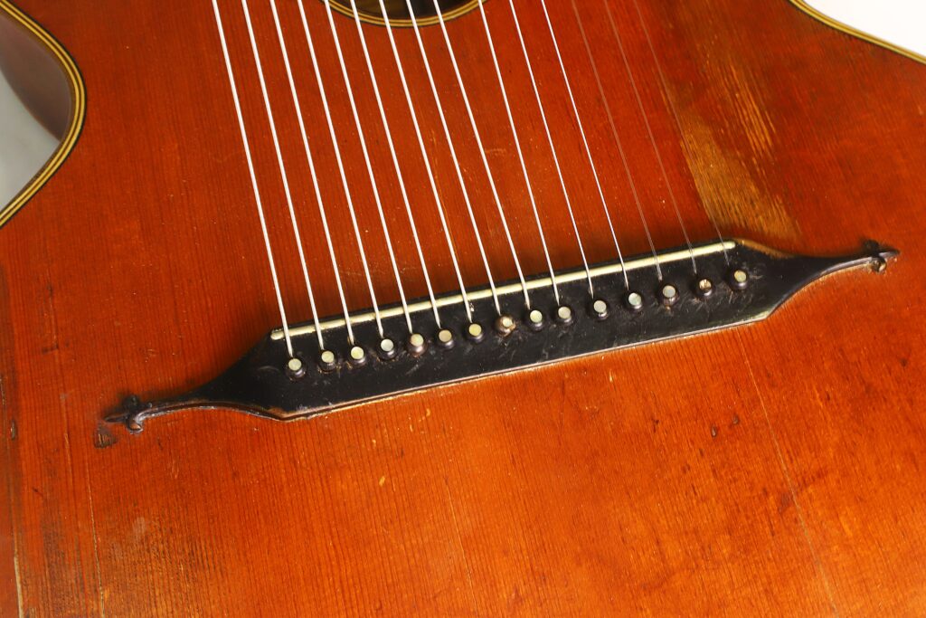 Nahaufnahme des Holzkörpers der „Gustav Keller Stauffer Stil 1900 Kontragitarre“ mit Fokus auf den Steg, die zahlreichen weißen Stifte und die Saiten. Die Oberfläche weist eine sichtbare Holzmaserung sowie einige Gebrauchsspuren auf. Nahaufnahme des Holzkörpers der „Gustav Keller Stauffer Stil 1900 Kontragitarre“ mit Fokus auf den Steg, die zahlreichen weißen Stifte und die Saiten. Die Oberfläche weist eine sichtbare Holzmaserung sowie einige Gebrauchsspuren auf.