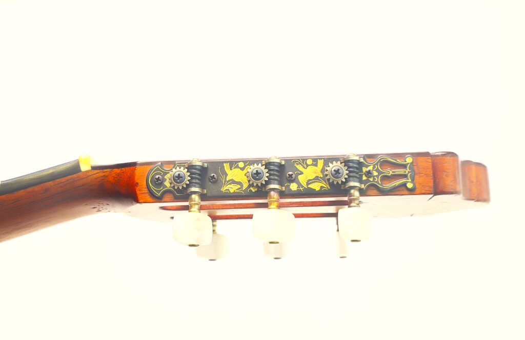 Nahaufnahme des Kopfes einer „Negra Flamenco“-Gitarre von Juan Lopez Aguilarte aus dem Jahr 1984, mit drei weißen Stimmwirbeln und einer kunstvoll verzierten Metall-Stimmplatte, die mit gelben Blumenmustern verziert ist und sich vor einem weißen Hintergrund abhebt. Nahaufnahme des Kopfes einer „Negra Flamenco“-Gitarre von Juan Lopez Aguilarte aus dem Jahr 1984, mit drei weißen Stimmwirbeln und einer kunstvoll verzierten Metall-Stimmplatte, die mit gelben Blumenmustern verziert ist und sich vor einem weißen Hintergrund abhebt.