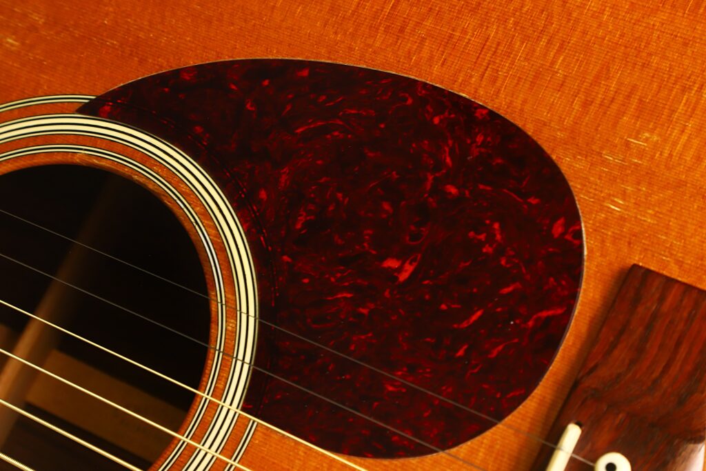 Nahaufnahme einer MARTIN OM-21 Akustikgitarre aus dem Jahr 1995, die einen Teil des Schalllochs, die Saiten, ein rotes Schlagbrett mit Schildpatt-Muster sowie die Holzmaserung des Gitarrenkorpus zeigt. Nahaufnahme einer MARTIN OM-21 Akustikgitarre aus dem Jahr 1995, die einen Teil des Schalllochs, die Saiten, ein rotes Schlagbrett mit Schildpatt-Muster sowie die Holzmaserung des Gitarrenkorpus zeigt.