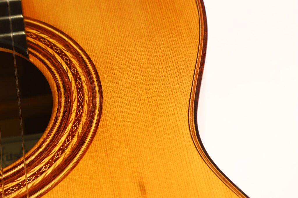 Nahaufnahme des Korpus der „Friederich Klassische Gitarre“ von Stefan Nitschke aus dem Jahr 2015 mit detaillierter Holzmaserung, einer kunstvollen Rosette um das Schallloch und einem Ausschnitt des Halses vor weißem Hintergrund. Nahaufnahme des Korpus der „Friederich Klassische Gitarre“ von Stefan Nitschke aus dem Jahr 2015 mit detaillierter Holzmaserung, einer kunstvollen Rosette um das Schallloch und einem Ausschnitt des Halses vor weißem Hintergrund.