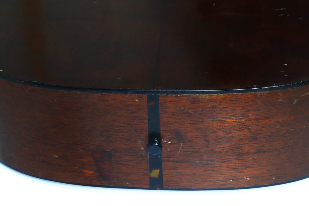 Nahaufnahme der Unterkante des Korpus einer Martin 000-18 Akustikgitarre aus dem Jahr 1960, auf der der Endstift und die Holzmaserung in der Lackierung zu sehen sind. Nahaufnahme der Unterkante des Korpus einer Martin 000-18 Akustikgitarre aus dem Jahr 1960, auf der der Endstift und die Holzmaserung in der Lackierung zu sehen sind.