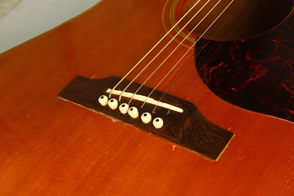 Nahaufnahme einer Gibson SJN 1968 Akustikgitarre, die den Steg aus Holz, die sechs Saiten, die weißen Stegstifte und einen Teil des Schlagbretts am Korpus zeigt. Nahaufnahme einer Gibson SJN 1968 Akustikgitarre, die den Steg aus Holz, die sechs Saiten, die weißen Stegstifte und einen Teil des Schlagbretts am Korpus zeigt.