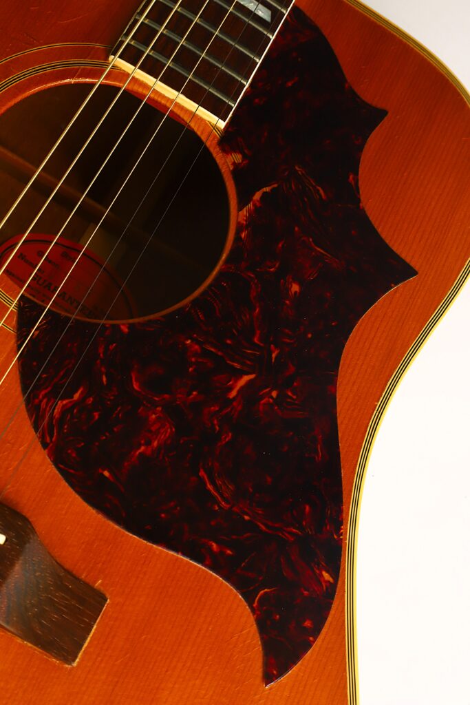 Nahaufnahme einer Gibson SJN 1968 Akustikgitarre, die die Saiten, das Schallloch, den Holzkorpus und ein großes, einzigartig geformtes, marmoriertes, dunkelrotes Schlagbrett zeigt. Das Bild hebt die Textur der Gitarre und ihre außergewöhnliche handwerkliche Verarbeitung hervor. Nahaufnahme einer Gibson SJN 1968 Akustikgitarre, die die Saiten, das Schallloch, den Holzkorpus und ein großes, einzigartig geformtes, marmoriertes, dunkelrotes Schlagbrett zeigt. Das Bild hebt die Textur der Gitarre und ihre außergewöhnliche handwerkliche Verarbeitung hervor.