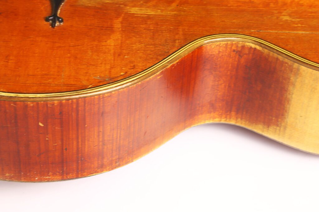Nahaufnahme der Seite und des Korpus der Kontragitarre „Gustav Keller Stauffer Stil 1900“ mit ihrer rotbraunen Holzmaserung, der goldenen Einfassung entlang der Kante und einigen Gebrauchsspuren. Der Hintergrund ist weiß. Nahaufnahme der Seite und des Korpus der Kontragitarre „Gustav Keller Stauffer Stil 1900“ mit ihrer rotbraunen Holzmaserung, der goldenen Einfassung entlang der Kante und einigen Gebrauchsspuren. Der Hintergrund ist weiß.
