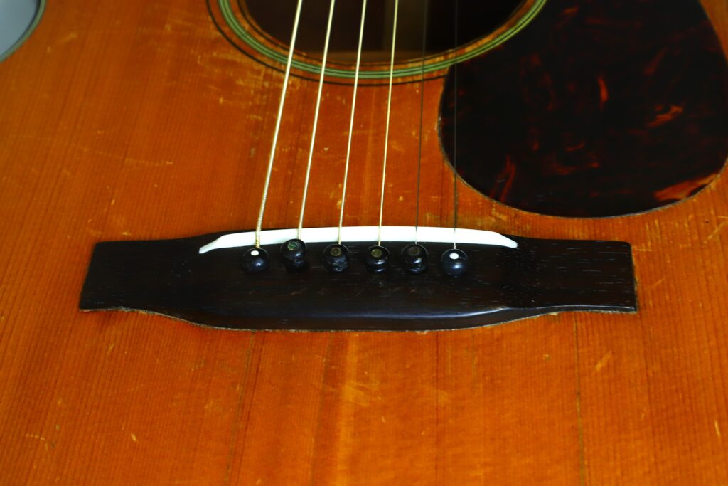 Nahaufnahme des Stegs der Martin 000-18 Akustikgitarre aus dem Jahr 1960, auf der sechs Saiten, schwarze Stegstifte und ein weißer Sattel auf dem Holzkorpus zu sehen sind. Nahaufnahme des Stegs der Martin 000-18 Akustikgitarre aus dem Jahr 1960, auf der sechs Saiten, schwarze Stegstifte und ein weißer Sattel auf dem Holzkorpus zu sehen sind.
