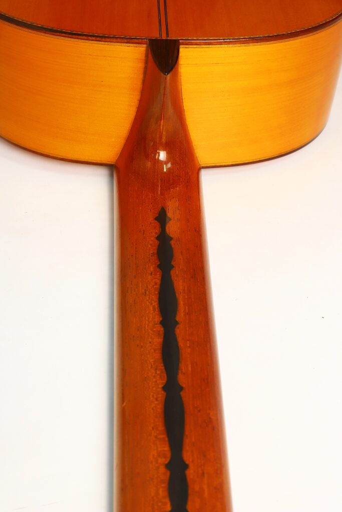 Nahaufnahme der Rückseite und des Halses der Flamenco-Gitarre „Jeronimo Pena 1976“, die den Hochglanzlack und die dunkle Zierintarsie in der Mitte des Halses hervorhebt. Nahaufnahme der Rückseite und des Halses der Flamenco-Gitarre „Jeronimo Pena 1976“, die den Hochglanzlack und die dunkle Zierintarsie in der Mitte des Halses hervorhebt.