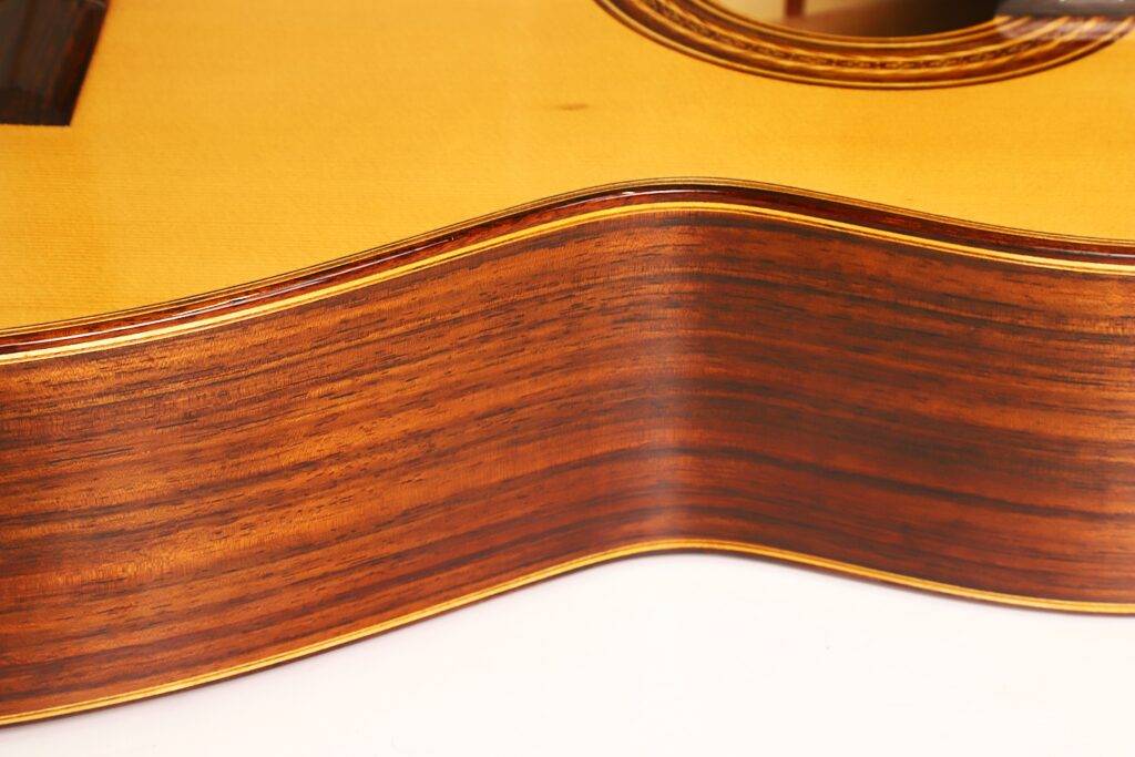 Nahaufnahme der gewölbten Seite einer „Friederich Klassische Gitarre“ von Stefan Nitschke aus dem Jahr 2015, die die Holzmaserung und die glänzende Lackierung der Gitarre zeigt, wobei im Hintergrund ein Teil des Schalllochs und des Korpus zu sehen ist. Nahaufnahme der gewölbten Seite einer „Friederich Klassische Gitarre“ von Stefan Nitschke aus dem Jahr 2015, die die Holzmaserung und die glänzende Lackierung der Gitarre zeigt, wobei im Hintergrund ein Teil des Schalllochs und des Korpus zu sehen ist.