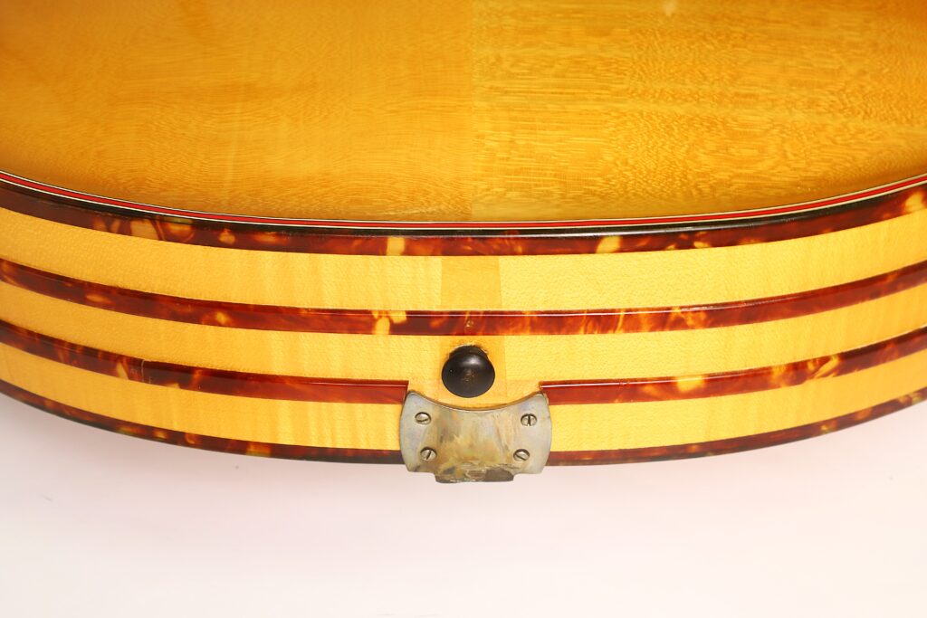Nahaufnahme der Seite einer Artur-Lang-Jazzgitarre aus den 1950er Jahren mit gelber Lackierung und dunkelbraunen Streifen, mit einem runden schwarzen Gurtknopf und einer Metallhalterung unten in der Mitte, vor weißem Hintergrund. Nahaufnahme der Seite einer Artur-Lang-Jazzgitarre aus den 1950er Jahren mit gelber Lackierung und dunkelbraunen Streifen, mit einem runden schwarzen Gurtknopf und einer Metallhalterung unten in der Mitte, vor weißem Hintergrund.