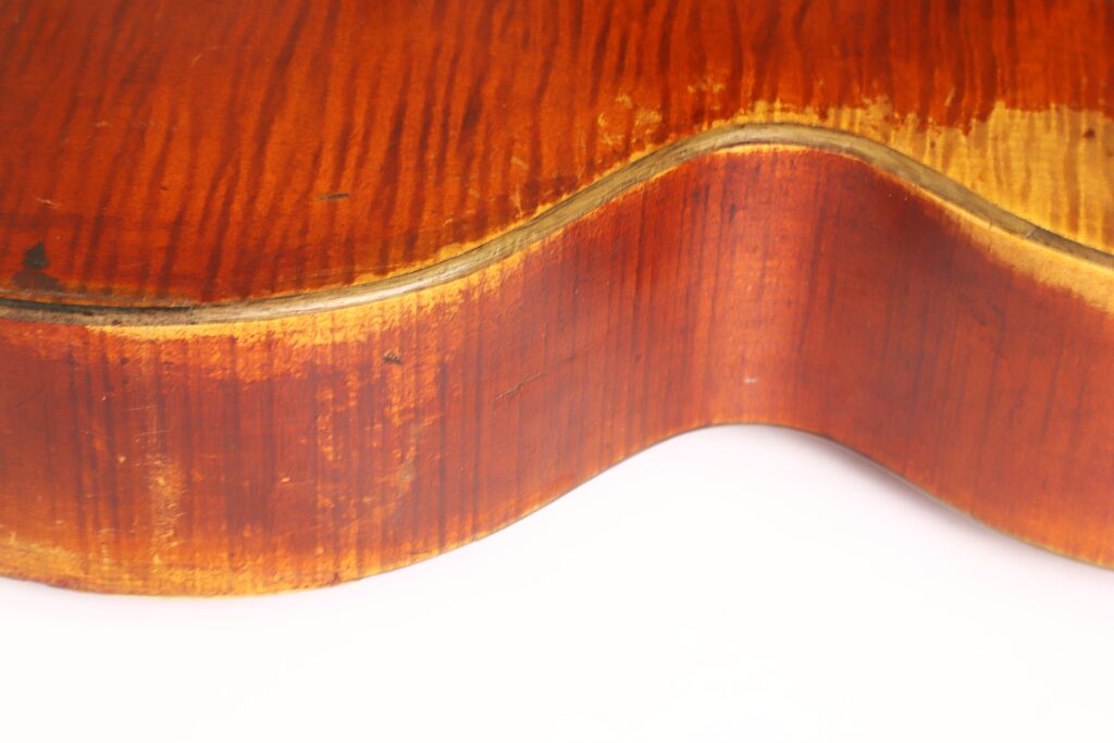 Nahaufnahme der Seite einer Kontragitarre vom Typ „Stil 1900“ von Gustav Keller Stauffer, die ihre geschwungene Form, die warme braune Farbe und die sichtbaren Maserungen des Holzes hervorhebt. Die glänzende Lackierung reflektiert das Licht, und einige Gebrauchsspuren unterstreichen ihren Vintage-Charakter. Nahaufnahme der Seite einer Kontragitarre vom Typ „Stil 1900“ von Gustav Keller Stauffer, die ihre geschwungene Form, die warme braune Farbe und die sichtbaren Maserungen des Holzes hervorhebt. Die glänzende Lackierung reflektiert das Licht, und einige Gebrauchsspuren unterstreichen ihren Vintage-Charakter.