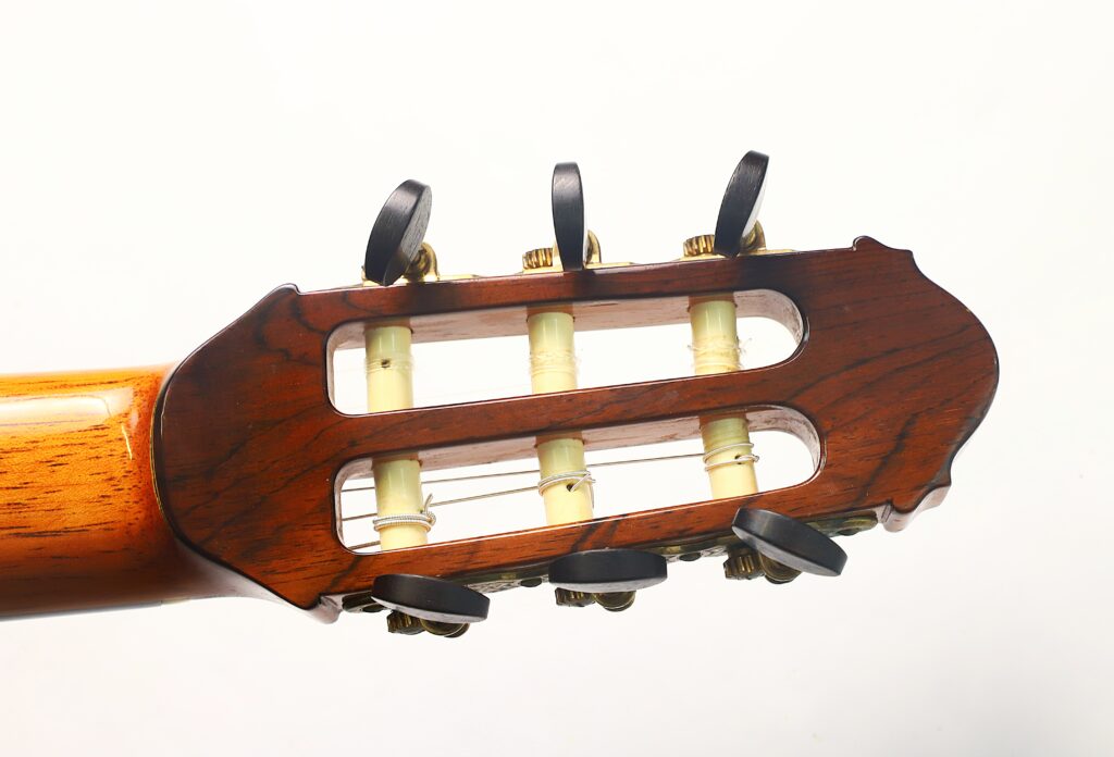 Nahaufnahme des Kopfstücks einer „Antonius Müller 2004 Doubletop Klassische Gitarre“, die die Holzstimmmechaniken und die weißen Stimmwirbel vor einem schlichten weißen Hintergrund zeigt. Nahaufnahme des Kopfstücks einer „Antonius Müller 2004 Doubletop Klassische Gitarre“, die die Holzstimmmechaniken und die weißen Stimmwirbel vor einem schlichten weißen Hintergrund zeigt.