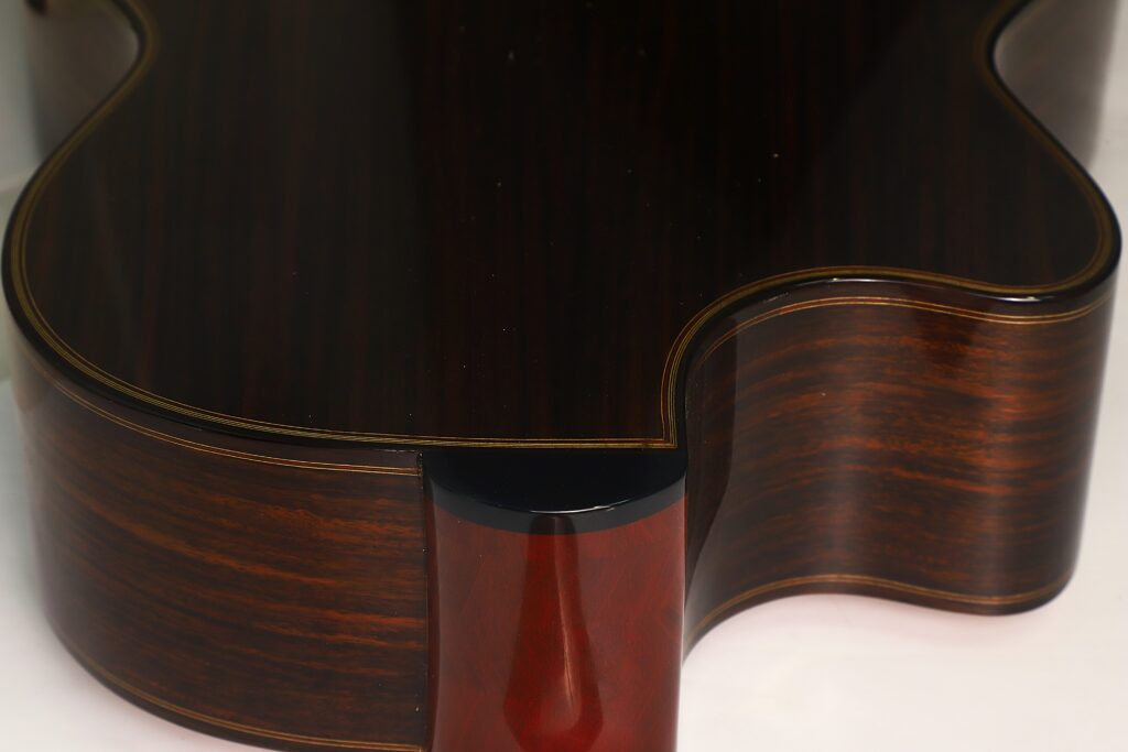 Nahaufnahme des Halsfußes und des Nackens einer „Doubletop Klassische Gitarre“ von Antonius Müller aus dem Jahr 2012, die die dunkle Holzmaserung, die glänzende Lackierung und die elegant geschwungene Kante vor einem hellen Hintergrund hervorhebt. Nahaufnahme des Halsfußes und des Nackens einer „Doubletop Klassische Gitarre“ von Antonius Müller aus dem Jahr 2012, die die dunkle Holzmaserung, die glänzende Lackierung und die elegant geschwungene Kante vor einem hellen Hintergrund hervorhebt.