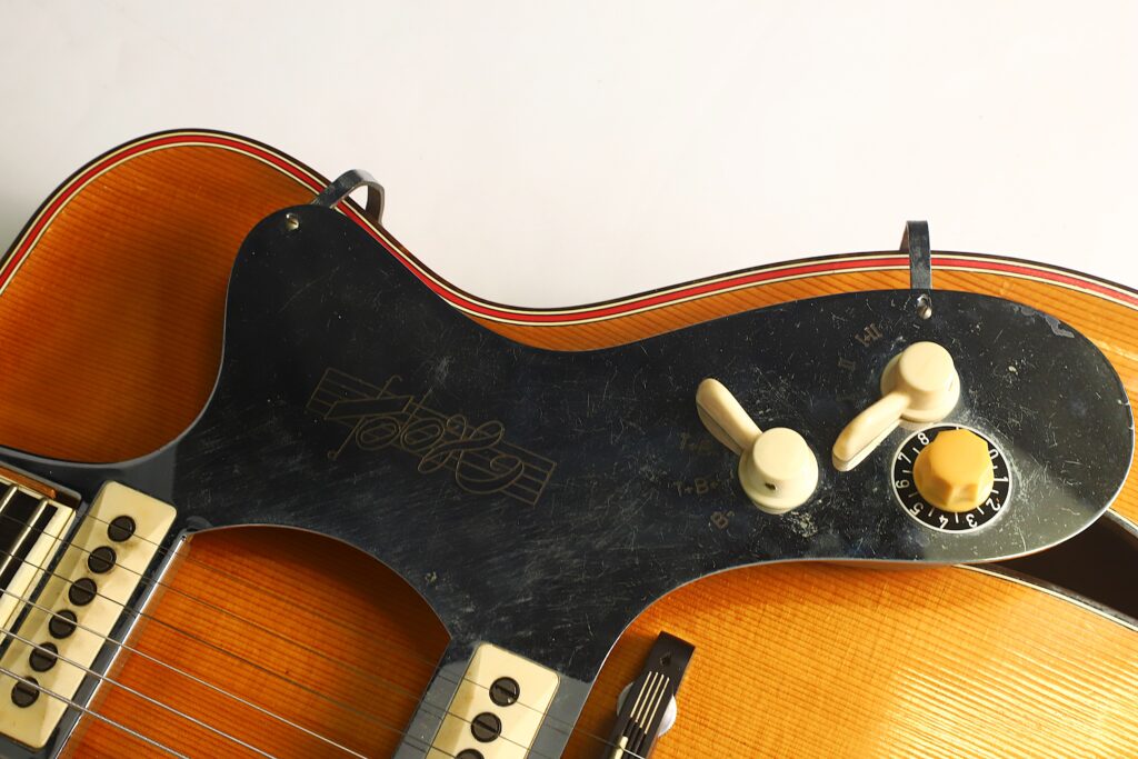 Nahaufnahme einer Artur Lang-E-Gitarre im Jazz-Stil aus den 1950er Jahren, bei der die Saiten, Tonabnehmer und das schwarze Schlagbrett mit zwei weißen und einem gelben Regler vor einem hellen Hintergrund im Vordergrund stehen. Nahaufnahme einer Artur Lang-E-Gitarre im Jazz-Stil aus den 1950er Jahren, bei der die Saiten, Tonabnehmer und das schwarze Schlagbrett mit zwei weißen und einem gelben Regler vor einem hellen Hintergrund im Vordergrund stehen.