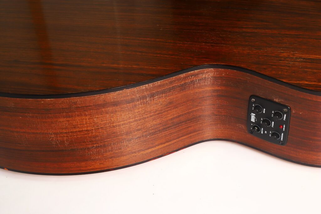 Nahaufnahme der Seite der MARTIN OM-21 Akustikgitarre aus dem Jahr 1995, die die integrierten elektronischen Bedienelemente und die charakteristische Holzmaserung hervorhebt. Die Gitarre zeichnet sich durch eine geschwungene Form und eine glänzende Lackierung aus. Nahaufnahme der Seite der MARTIN OM-21 Akustikgitarre aus dem Jahr 1995, die die integrierten elektronischen Bedienelemente und die charakteristische Holzmaserung hervorhebt. Die Gitarre zeichnet sich durch eine geschwungene Form und eine glänzende Lackierung aus.