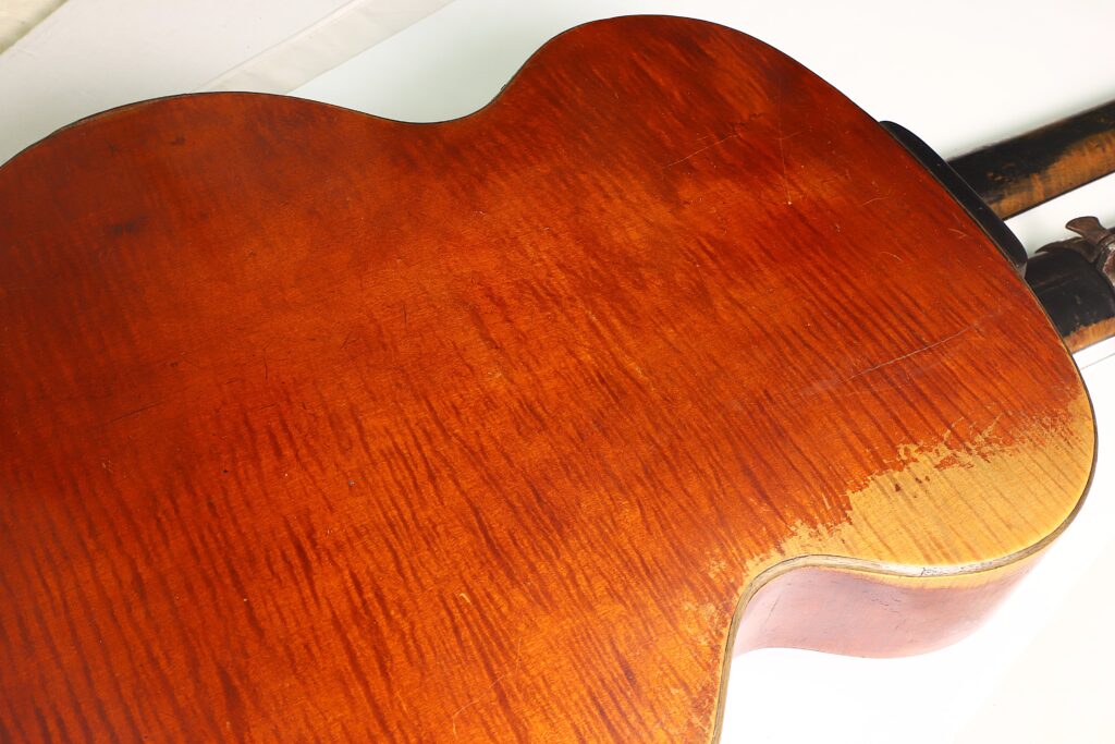 Nahaufnahme der Rückseite der Kontragitarre „Gustav Keller Stauffer Stil 1900“ mit einer glänzenden, rotbraunen Holzoberfläche. Die Holzmaserung ist sichtbar, am unteren rechten Rand sind einige Gebrauchsspuren und Verfärbungen zu erkennen. Nahaufnahme der Rückseite der Kontragitarre „Gustav Keller Stauffer Stil 1900“ mit einer glänzenden, rotbraunen Holzoberfläche. Die Holzmaserung ist sichtbar, am unteren rechten Rand sind einige Gebrauchsspuren und Verfärbungen zu erkennen.