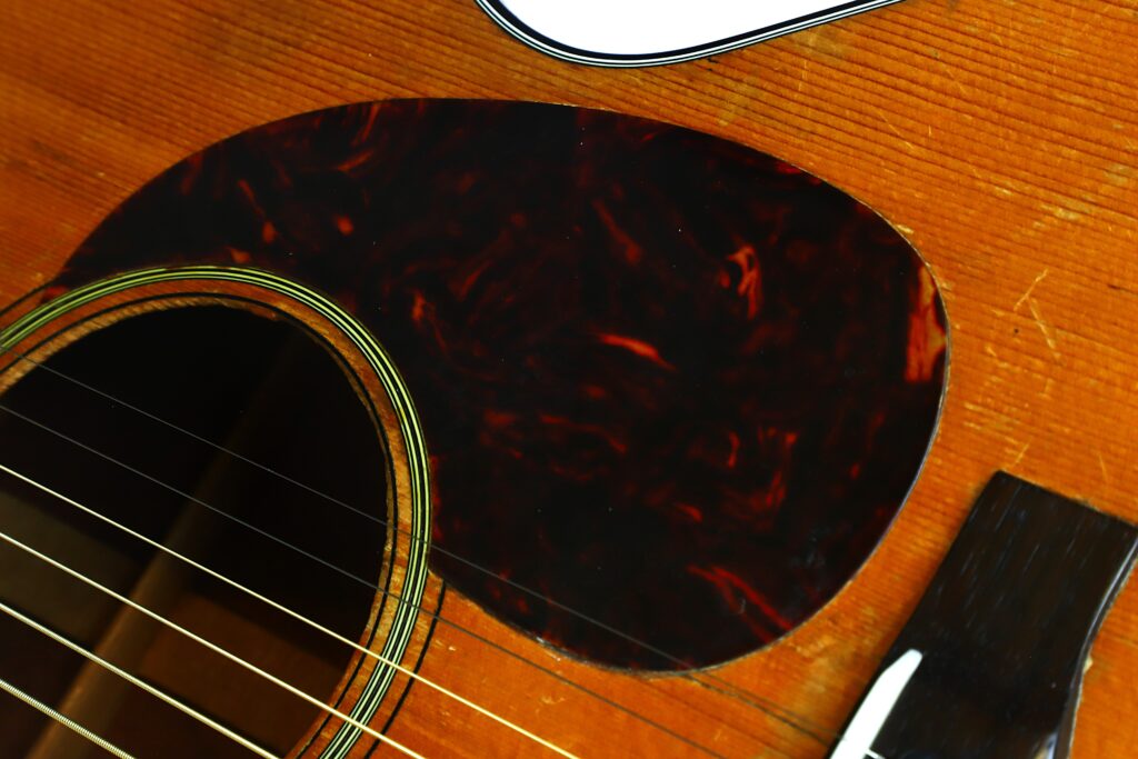 Nahaufnahme der Martin 000-18 Akustikgitarre aus dem Jahr 1960, bei der das Schallloch, die Saiten, das Schlagbrett mit Schildpatt-Muster und ein Teil des Holzstegs besonders hervorgehoben sind. Nahaufnahme der Martin 000-18 Akustikgitarre aus dem Jahr 1960, bei der das Schallloch, die Saiten, das Schlagbrett mit Schildpatt-Muster und ein Teil des Holzstegs besonders hervorgehoben sind.
