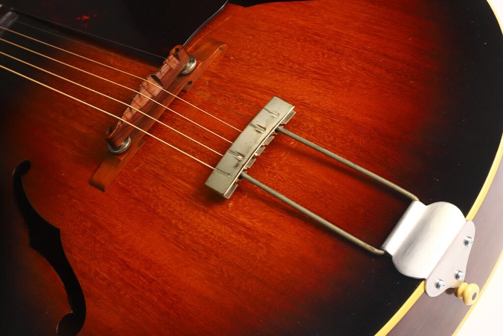 Nahaufnahme des Korpus einer Gibson L-48 Akustikgitarre aus dem Jahr 1948, bei der die Saiten, der Holzsteg, der Metall-Saitenhalter und die Sunburst-Lackierung besonders hervorgehoben sind. Nahaufnahme des Korpus einer Gibson L-48 Akustikgitarre aus dem Jahr 1948, bei der die Saiten, der Holzsteg, der Metall-Saitenhalter und die Sunburst-Lackierung besonders hervorgehoben sind.