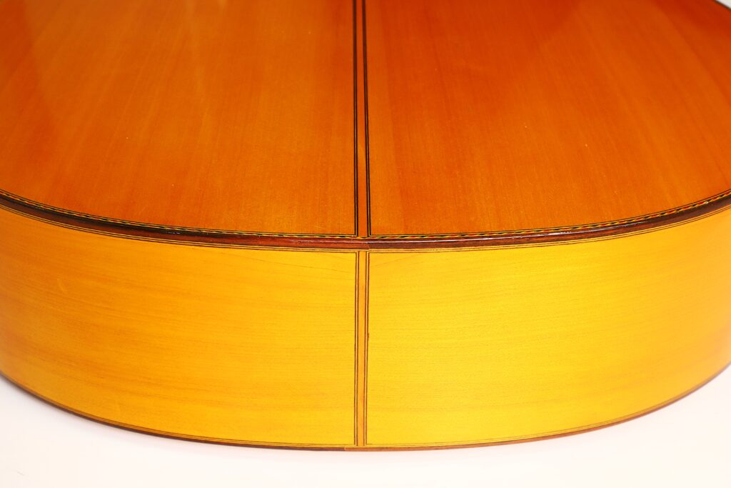 Nahaufnahme der Rückseite und der Zargen der Flamenco-Gitarre „Jeronimo Pena 1976“, die die Holzmaserung, die Hochglanzlackierung und die mittige Fuge zwischen den beiden Holzstücken hervorhebt. Nahaufnahme der Rückseite und der Zargen der Flamenco-Gitarre „Jeronimo Pena 1976“, die die Holzmaserung, die Hochglanzlackierung und die mittige Fuge zwischen den beiden Holzstücken hervorhebt.