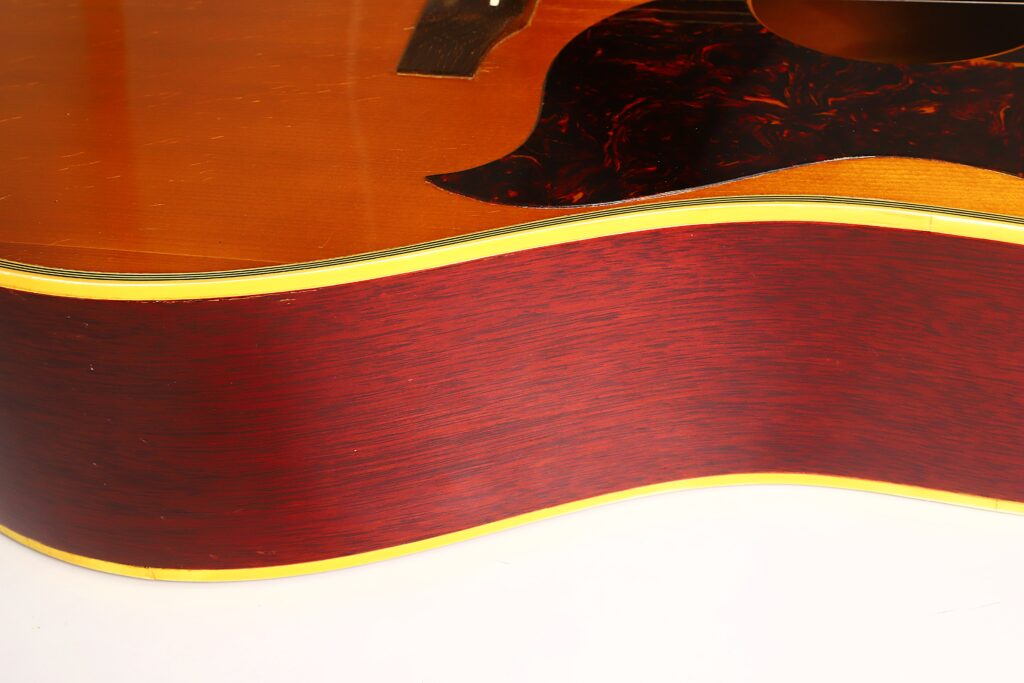 Nahaufnahme der Gibson SJN 1968 Akustikgitarre, die die rotbraune Holzmaserung an den Zargen und am Korpus, die gelbe Einfassung sowie einen Teil des dunklen Schildpatt-Schlagbretts hervorhebt. Nahaufnahme der Gibson SJN 1968 Akustikgitarre, die die rotbraune Holzmaserung an den Zargen und am Korpus, die gelbe Einfassung sowie einen Teil des dunklen Schildpatt-Schlagbretts hervorhebt.