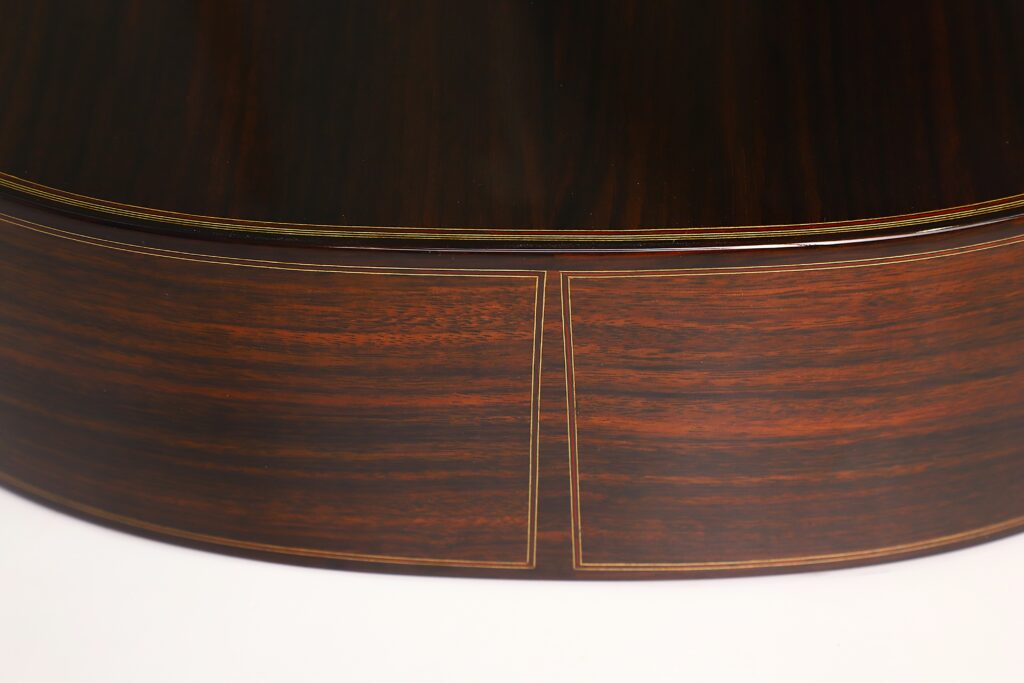 Nahaufnahme der Rückseite und der Seite der „Antonius Müller 2012 Doubletop Klassische Gitarre“, die das dunkle, polierte Holz mit seiner feinen Maserung und den dekorativen Einlegearbeiten hervorhebt. Nahaufnahme der Rückseite und der Seite der „Antonius Müller 2012 Doubletop Klassische Gitarre“, die das dunkle, polierte Holz mit seiner feinen Maserung und den dekorativen Einlegearbeiten hervorhebt.