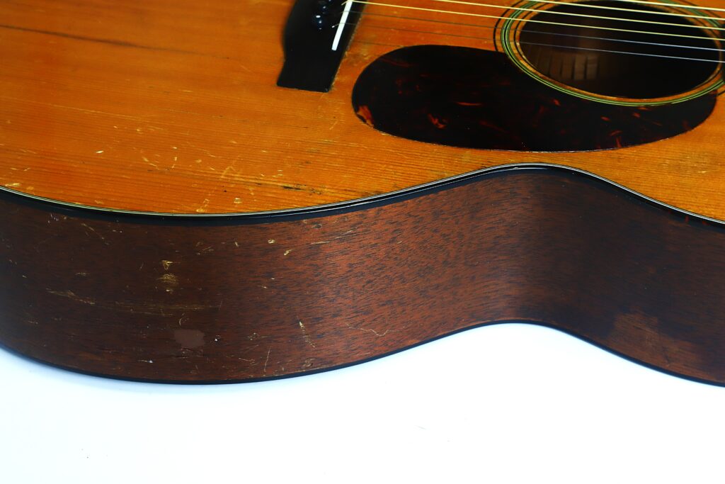Nahaufnahme des Korpus einer Martin 000-18 Akustikgitarre aus dem Jahr 1960, die die Holzmaserung, die Saiten, einen Teil des Schalllochs und einige Kratzer auf der Oberfläche zeigt, vor einem weißen Hintergrund. Nahaufnahme des Korpus einer Martin 000-18 Akustikgitarre aus dem Jahr 1960, die die Holzmaserung, die Saiten, einen Teil des Schalllochs und einige Kratzer auf der Oberfläche zeigt, vor einem weißen Hintergrund.