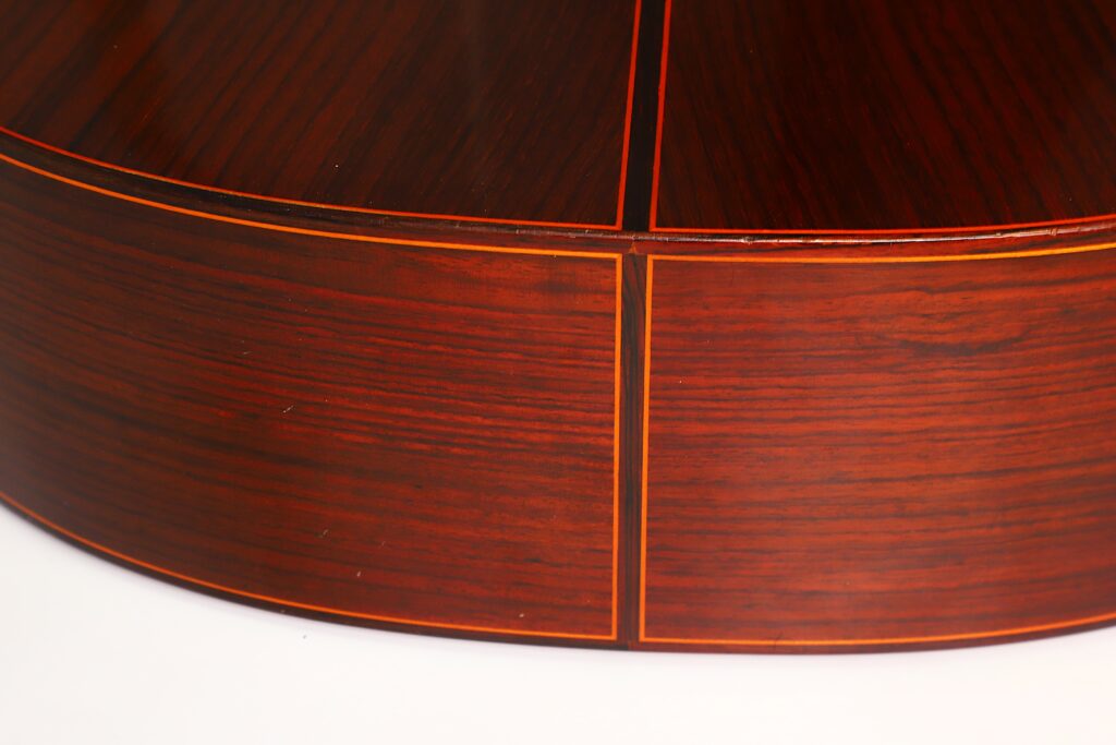 Nahaufnahme der polierten Holzoberfläche einer Jose Ramirez 1a 1977 Klassische Gitarre, die sich durch dunkle und helle rotbraune Maserungen auszeichnet, ergänzt durch schmale, helle Einlegearbeiten, die geometrische Formen bilden. Nahaufnahme der polierten Holzoberfläche einer Jose Ramirez 1a 1977 Klassische Gitarre, die sich durch dunkle und helle rotbraune Maserungen auszeichnet, ergänzt durch schmale, helle Einlegearbeiten, die geometrische Formen bilden.
