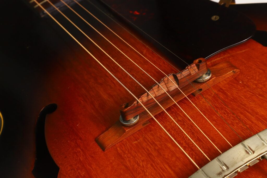Nahaufnahme der Holzbrücke und der Saiten einer Gibson L-48 Akustikgitarre aus dem Jahr 1948, die die feinen Details des braunen Korpus und der Beschläge der Gitarre hervorhebt. Nahaufnahme der Holzbrücke und der Saiten einer Gibson L-48 Akustikgitarre aus dem Jahr 1948, die die feinen Details des braunen Korpus und der Beschläge der Gitarre hervorhebt.