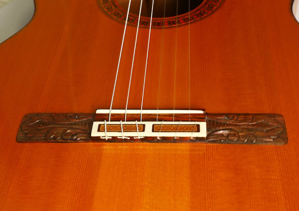 Nahaufnahme des Korpus der Flamenco-Gitarre „Jeronimo Pena 1976“, bei der die Saiten und der aufwendig gearbeitete Holzsteg vor dem Hintergrund einer warmen braunen Lackierung besonders gut zur Geltung kommen. Nahaufnahme des Korpus der Flamenco-Gitarre „Jeronimo Pena 1976“, bei der die Saiten und der aufwendig gearbeitete Holzsteg vor dem Hintergrund einer warmen braunen Lackierung besonders gut zur Geltung kommen.
