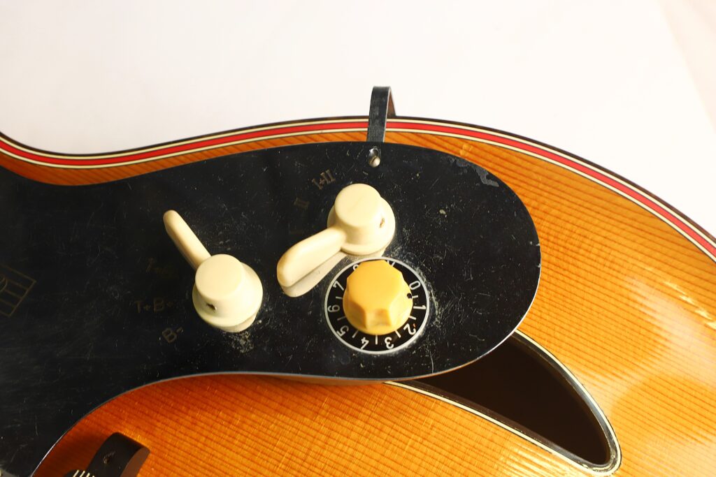 Nahaufnahme einer elektrischen Jazz-Gitarre von Artur Lang aus den 1950er Jahren, auf der zwei weiße Regler und ein gelber Regler mit den Zahlen 1 bis 12 vor einem schwarzen Schlagbrett zu sehen sind, wobei ein Teil des hölzernen Gitarrenkorpus sichtbar ist. Nahaufnahme einer elektrischen Jazz-Gitarre von Artur Lang aus den 1950er Jahren, auf der zwei weiße Regler und ein gelber Regler mit den Zahlen 1 bis 12 vor einem schwarzen Schlagbrett zu sehen sind, wobei ein Teil des hölzernen Gitarrenkorpus sichtbar ist.