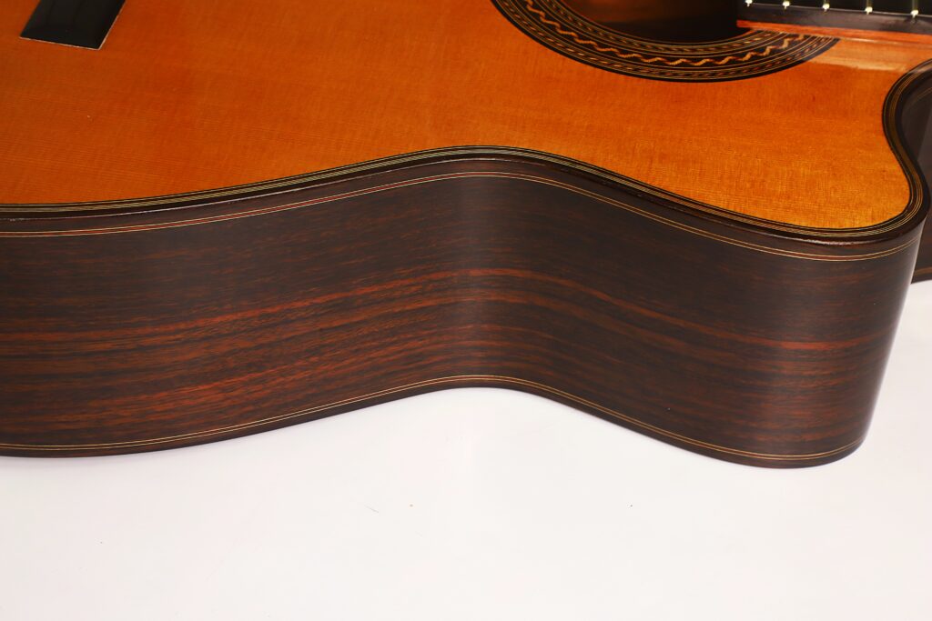 Nahaufnahme der Seite und des Korpus der „Antonius Müller 2012 Doubletop Klassische Gitarre“, die die dunkle Holzmaserung und die detailreiche Handwerkskunst vor weißem Hintergrund hervorhebt. Nahaufnahme der Seite und des Korpus der „Antonius Müller 2012 Doubletop Klassische Gitarre“, die die dunkle Holzmaserung und die detailreiche Handwerkskunst vor weißem Hintergrund hervorhebt.