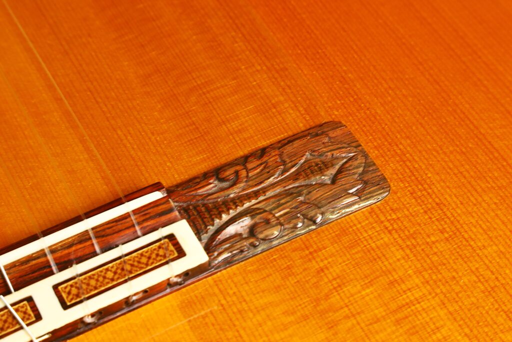 Nahaufnahme des Stegs der Flamenco-Gitarre „Jeronimo Pena 1976“ mit aufwendigen Holzschnitzereien und weißen Einlagen auf einem polierten, goldbraunen Holzkorpus. Nahaufnahme des Stegs der Flamenco-Gitarre „Jeronimo Pena 1976“ mit aufwendigen Holzschnitzereien und weißen Einlagen auf einem polierten, goldbraunen Holzkorpus.