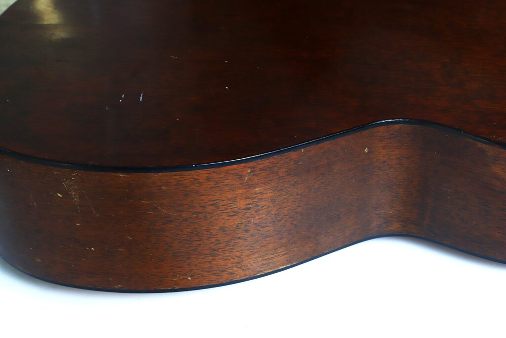 Nahaufnahme der Seite und Rückseite einer Martin 000-18 Akustikgitarre aus dem Jahr 1960, die ihre dunkelbraune, polierte Holzoberfläche mit leichten Kratzern zeigt. Die Gitarre liegt auf einer weißen Unterlage. Nahaufnahme der Seite und Rückseite einer Martin 000-18 Akustikgitarre aus dem Jahr 1960, die ihre dunkelbraune, polierte Holzoberfläche mit leichten Kratzern zeigt. Die Gitarre liegt auf einer weißen Unterlage.