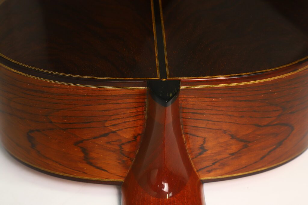 Nahaufnahme des Hals-Korpus-Übergangs der „Antonius Müller 2004 Doubletop Klassische Gitarre“, die die Holzmaserung, die glänzende Lackierung und die feinen Einlegearbeiten zeigt. Das Bild unterstreicht die handwerkliche Kunstfertigkeit und die hervorragende Materialqualität dieser klassischen Gitarre. Nahaufnahme des Hals-Korpus-Übergangs der „Antonius Müller 2004 Doubletop Klassische Gitarre“, die die Holzmaserung, die glänzende Lackierung und die feinen Einlegearbeiten zeigt. Das Bild unterstreicht die handwerkliche Kunstfertigkeit und die hervorragende Materialqualität dieser klassischen Gitarre.