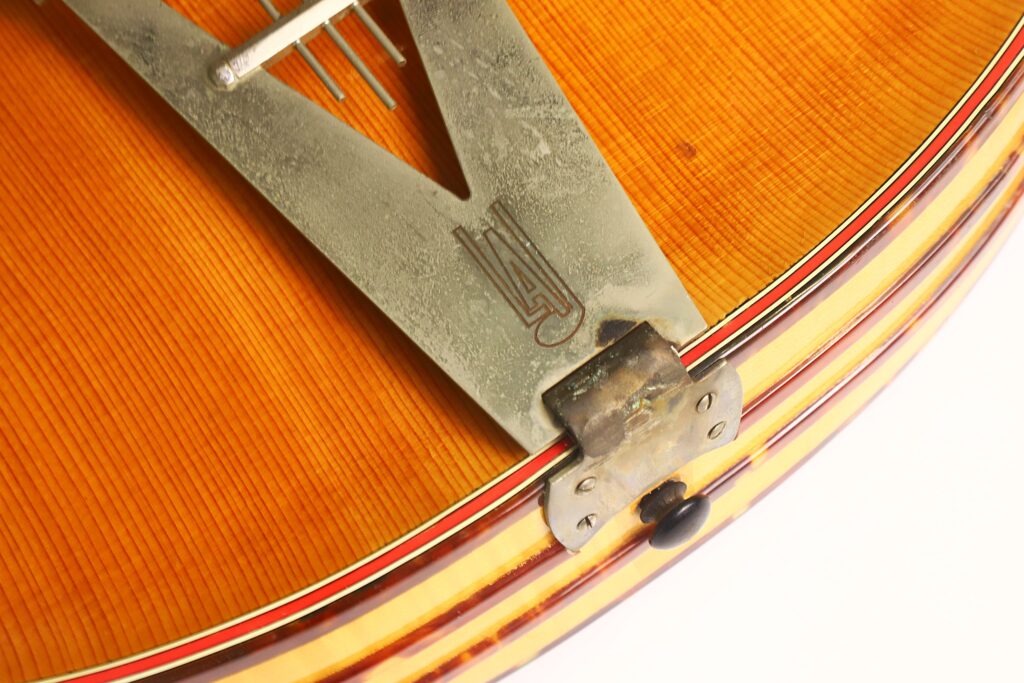 Nahaufnahme des Metall-Saitenhalters, der am Holzkorpus einer Artur-Lang-Jazzgitarre aus den 1950er Jahren befestigt ist; dabei kommen die feine Holzmaserung und die mehrschichtige Einfassung besonders gut zur Geltung. Nahaufnahme des Metall-Saitenhalters, der am Holzkorpus einer Artur-Lang-Jazzgitarre aus den 1950er Jahren befestigt ist; dabei kommen die feine Holzmaserung und die mehrschichtige Einfassung besonders gut zur Geltung.