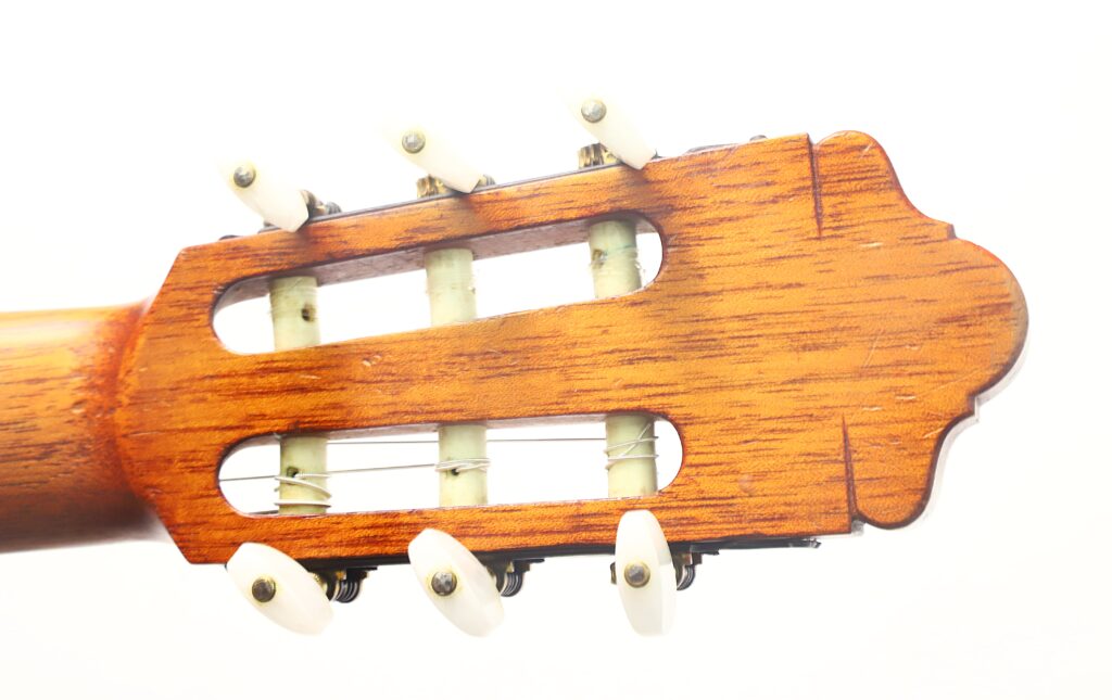 Nahaufnahme des hölzernen Kopfstücks der „Juan Lopez Aguilarte 1984 Negra Flamenco“-Gitarre, auf der weiße Stimmwirbel und Saiten vor weißem Hintergrund zu sehen sind. Nahaufnahme des hölzernen Kopfstücks der „Juan Lopez Aguilarte 1984 Negra Flamenco“-Gitarre, auf der weiße Stimmwirbel und Saiten vor weißem Hintergrund zu sehen sind.
