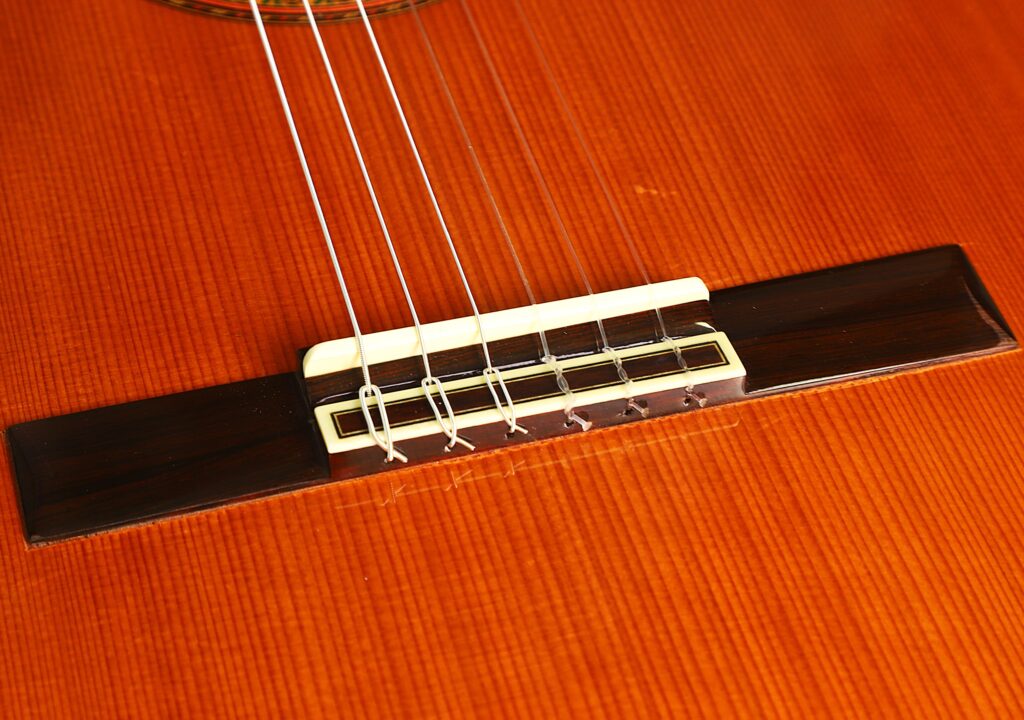 Eine detaillierte Nahaufnahme des Stegs und der Saiten der „Gran Klassische Gitarre“ von Raimundo y Aparicio Corujo vor dem Hintergrund ihres polierten Holzkörpers, die die aufwendige Handwerkskunst des Instruments verdeutlicht. Eine detaillierte Nahaufnahme des Stegs und der Saiten der „Gran Klassische Gitarre“ von Raimundo y Aparicio Corujo vor dem Hintergrund ihres polierten Holzkörpers, die die aufwendige Handwerkskunst des Instruments verdeutlicht.