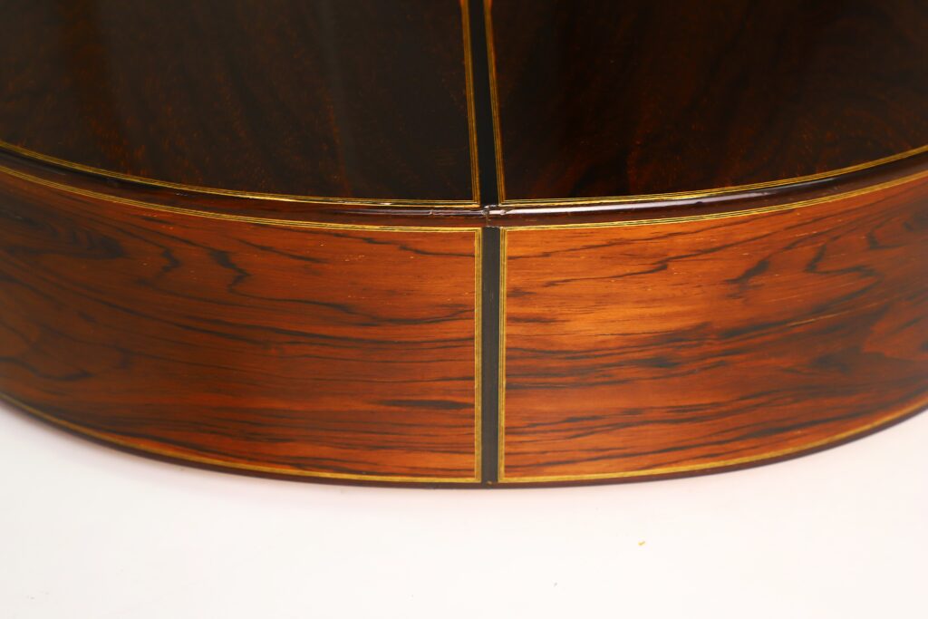 Nahaufnahme der polierten Holzdecke der „Antonius Müller 2004 Doubletop Klassische Gitarre“, die aufwendige Maserungsmuster zeigt, die durch feine goldfarbene Linien vor einem weißen Hintergrund hervorgehoben werden. Nahaufnahme der polierten Holzdecke der „Antonius Müller 2004 Doubletop Klassische Gitarre“, die aufwendige Maserungsmuster zeigt, die durch feine goldfarbene Linien vor einem weißen Hintergrund hervorgehoben werden.