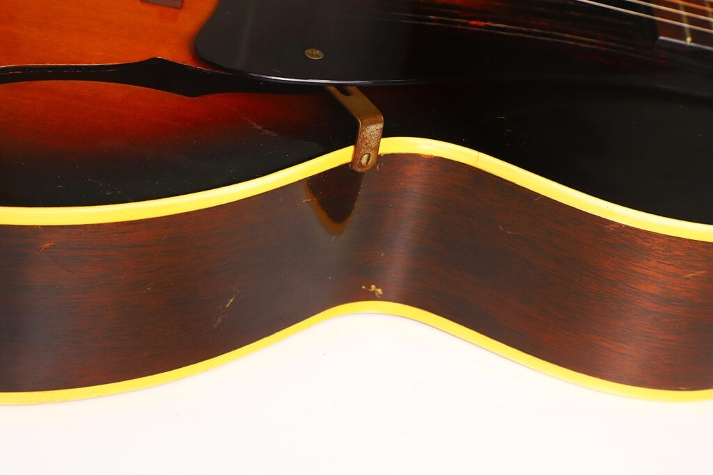 Nahaufnahme der Seite einer Gibson L-48 Akustikgitarre aus dem Jahr 1948 mit Sunburst-Lackierung, gelber Einfassung und einer kleinen Metallhalterung nahe der Oberkante. Nahaufnahme der Seite einer Gibson L-48 Akustikgitarre aus dem Jahr 1948 mit Sunburst-Lackierung, gelber Einfassung und einer kleinen Metallhalterung nahe der Oberkante.