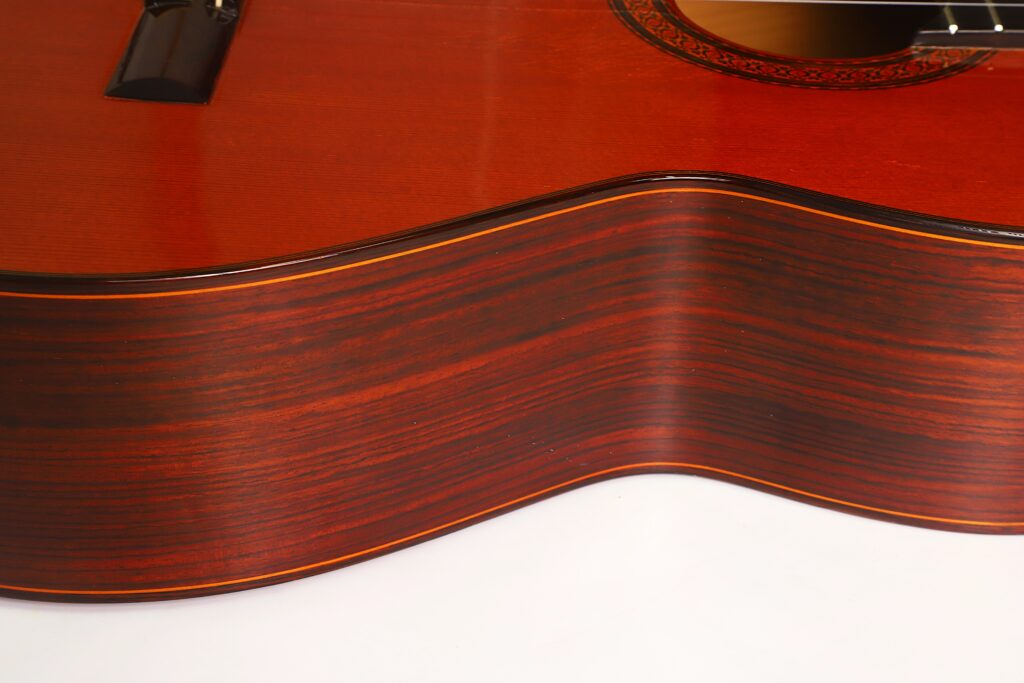 Nahaufnahme der Seite einer „Jose Ramirez 1a 1977 Klassische Gitarre“, die die polierte Holzoberfläche und die markante Maserung hervorhebt; dabei sind ein Teil der Gitarrendecke und das dekorative Schallloch zu sehen. Nahaufnahme der Seite einer „Jose Ramirez 1a 1977 Klassische Gitarre“, die die polierte Holzoberfläche und die markante Maserung hervorhebt; dabei sind ein Teil der Gitarrendecke und das dekorative Schallloch zu sehen.