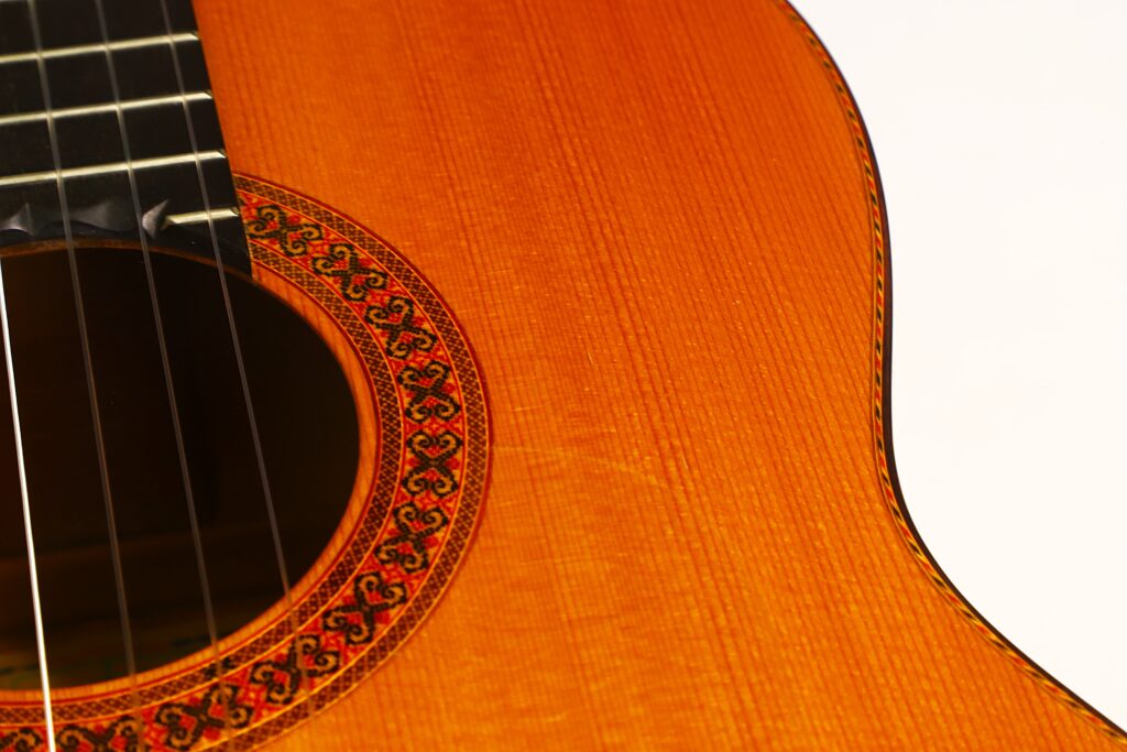 Nahaufnahme des Korpus der Flamenco-Gitarre „Jeronimo Pena 1976“, bei der das kunstvoll verzierte Schallloch, die Saiten und die feine Holzmaserung hervorgehoben werden, wobei ein Teil der Gitarre vor einem weißen Hintergrund zu sehen ist. Nahaufnahme des Korpus der Flamenco-Gitarre „Jeronimo Pena 1976“, bei der das kunstvoll verzierte Schallloch, die Saiten und die feine Holzmaserung hervorgehoben werden, wobei ein Teil der Gitarre vor einem weißen Hintergrund zu sehen ist.